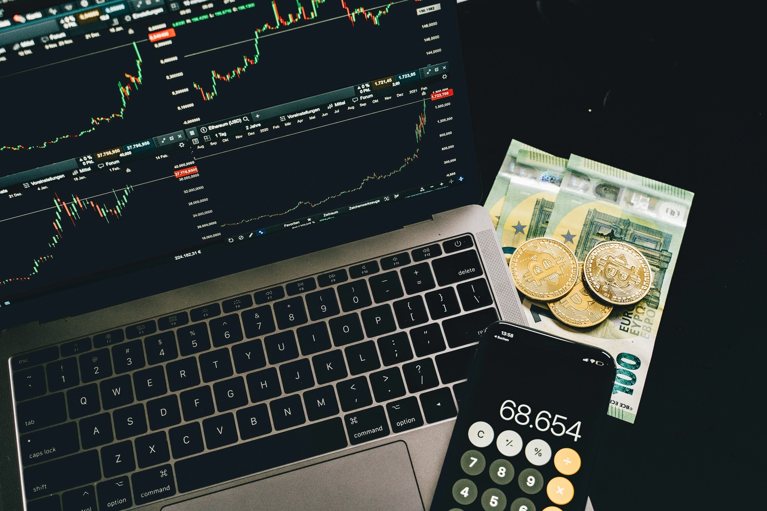 BIM - Bayerisches InvestManagement - InvestManagement -  Ein Laptop mit Finanzmarktdiagrammen, ein Taschenrechner mit der Zahl 68.654, Goldmünzen und Euro-Banknoten deuten auf Kryptowährungshandel oder Finanzanalyse hin.