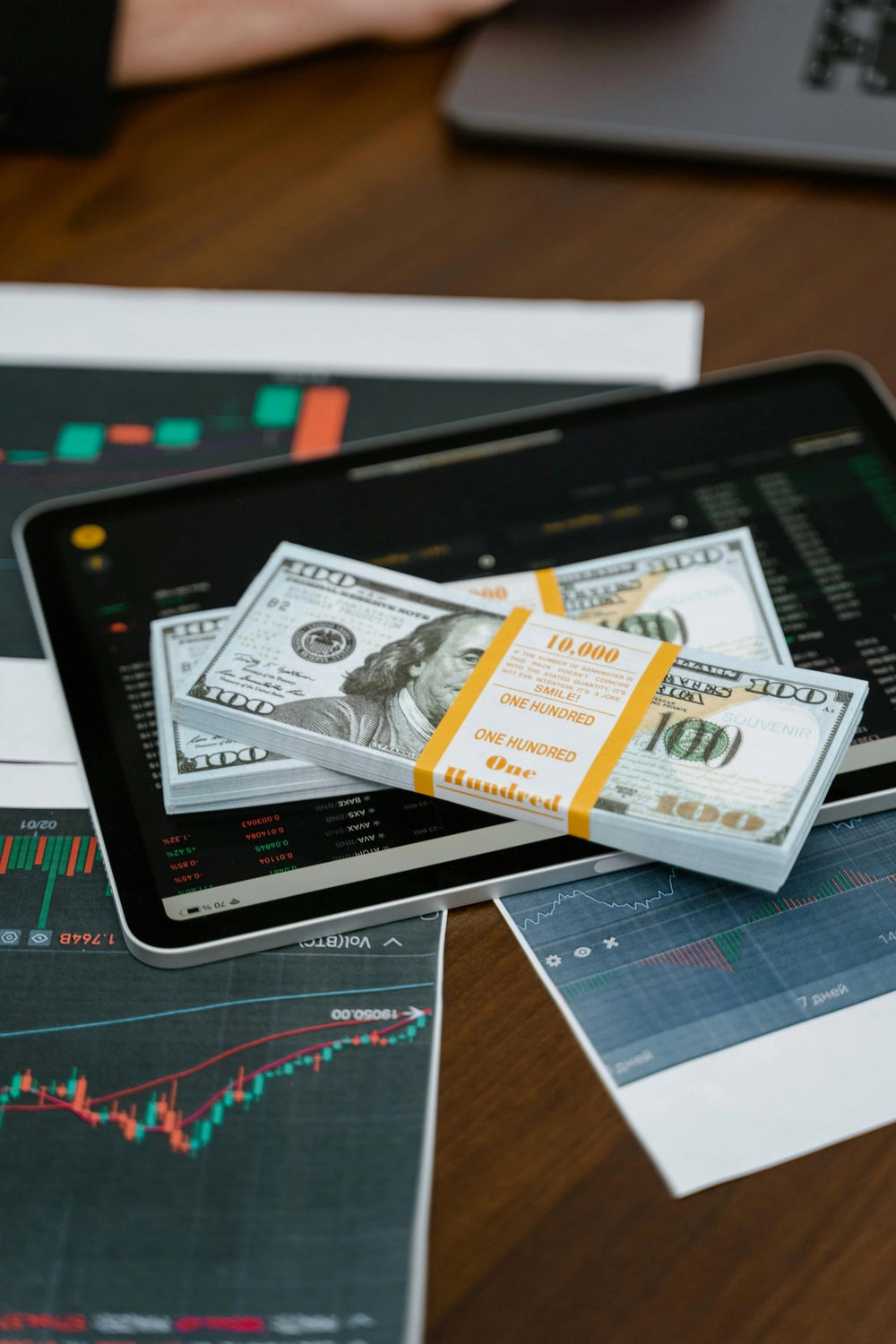 BIM - Bayerisches InvestManagement - InvestManagement -  Stapel von US-Hundert-Dollar-Scheinen lagen auf einem Laptop, auf dem Finanzmarktdiagramme und -daten angezeigt wurden.