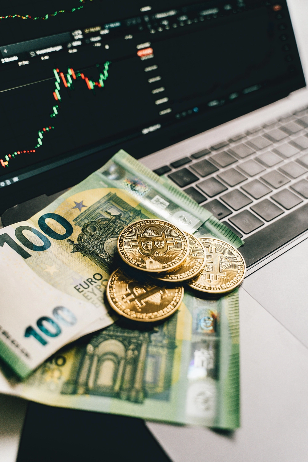 BIM - Bayerisches InvestManagement - InvestManagement -  Eine 100-Euro-Banknote und goldfarbene Münzen, die wahrscheinlich Bitcoin repräsentieren, liegen auf einer Laptop-Tastatur, auf deren Bildschirm ein Finanzhandelsdiagramm angezeigt wird.